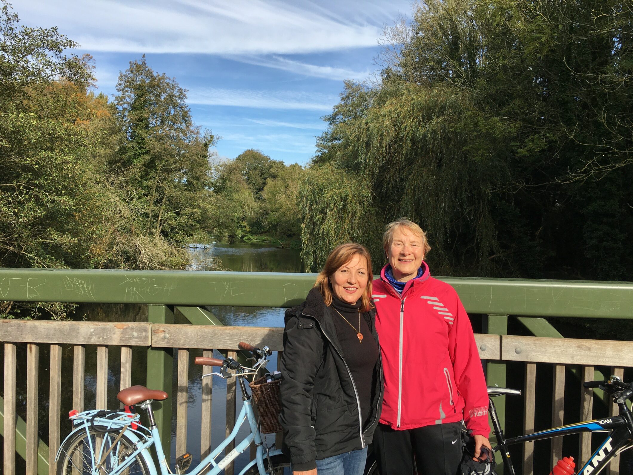 Two women standing next to each other on a bridge smiling at the camera, with their bikes on a bridge
