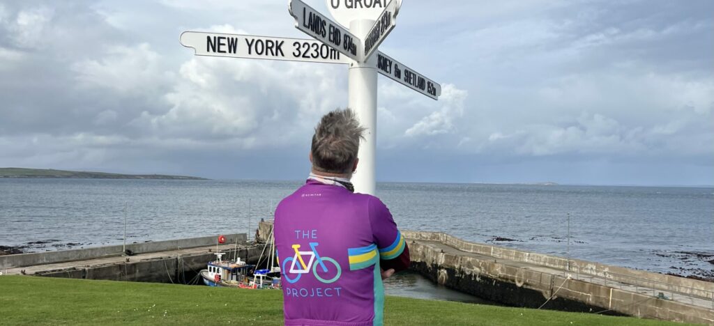 A man in a purple cycling jersey admiring the John O'Groats roadsign at the tip of the UK