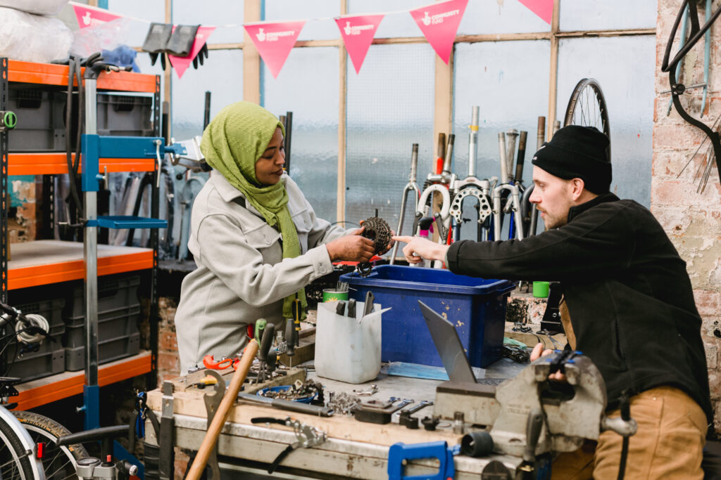 A mechanic in a bike workshop teaches a student how to fix a bicycle gear