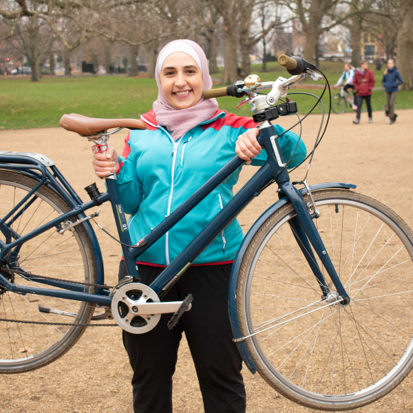 A woman in a colourful outfit and hijab, smiling and confidently holding up her bike in a park