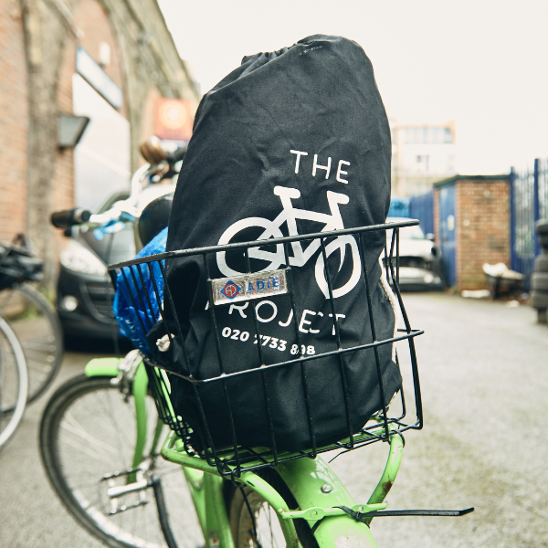 A black drawstring bag with The Bike Project's logo in a basket on the back of a green bike