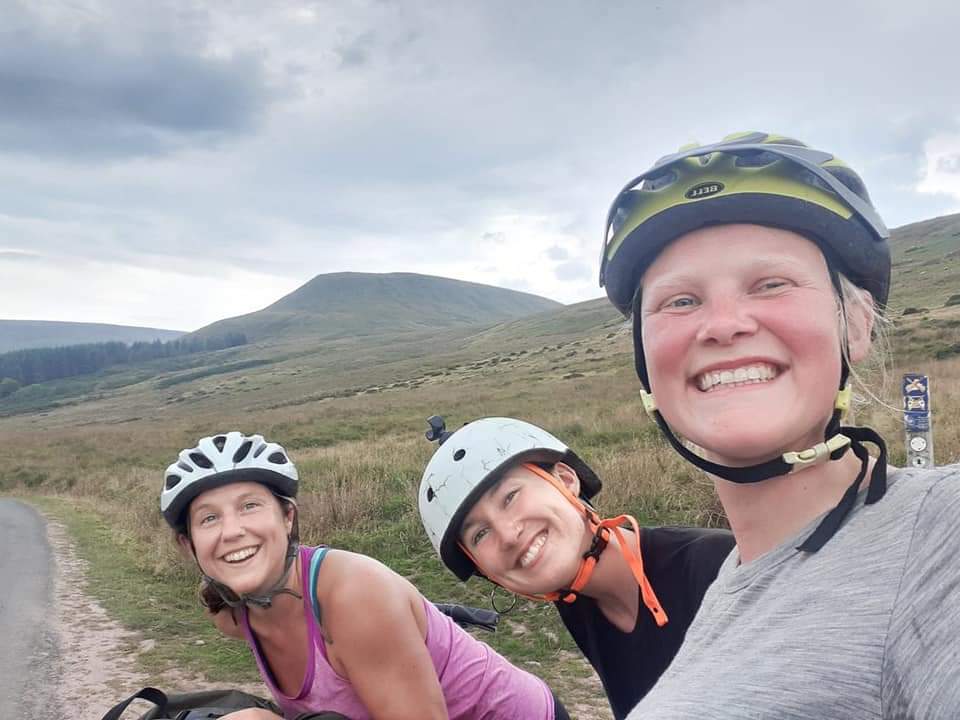 Three women cyclists smiling and taking a selfie wearing cycling helmets on an open countryside road