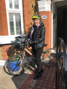 A woman in a black coat and cycling helmet posing with a bike in front of a house